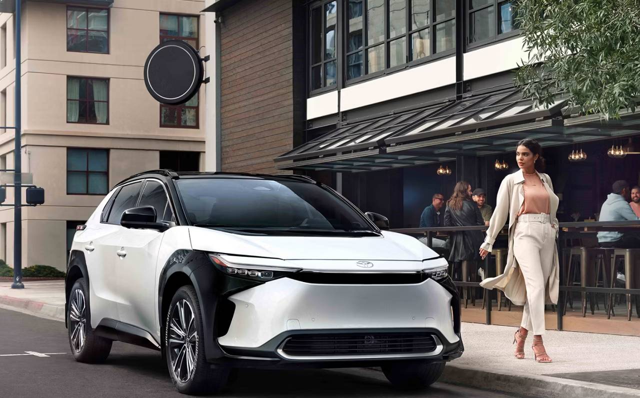 A woman in a fashionable white suit walks alongside a white and black 2025 Bz4X outside an upscale cafe