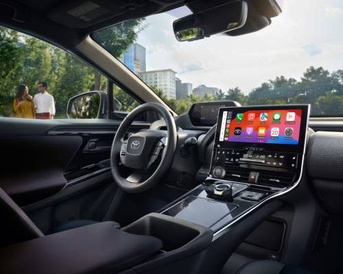 The steering wheel and dashboard display inside the 2025 Toyota Bz4X. Outside the front and driver's window, people walk by on a pleasantly sunny day.