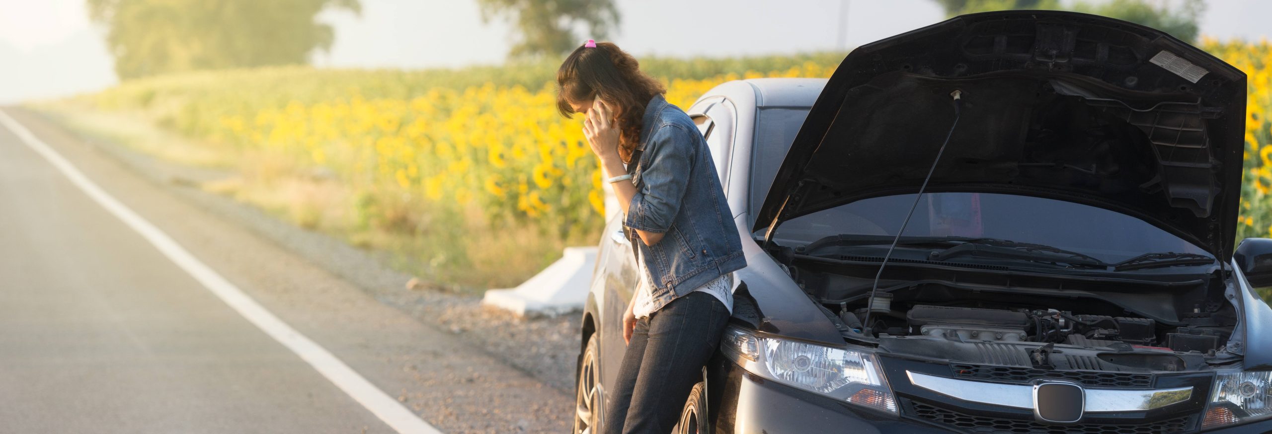 A woman is having a phone call beside her car which has broken down on the side of a flower-lined road. The hood of her car is popped.