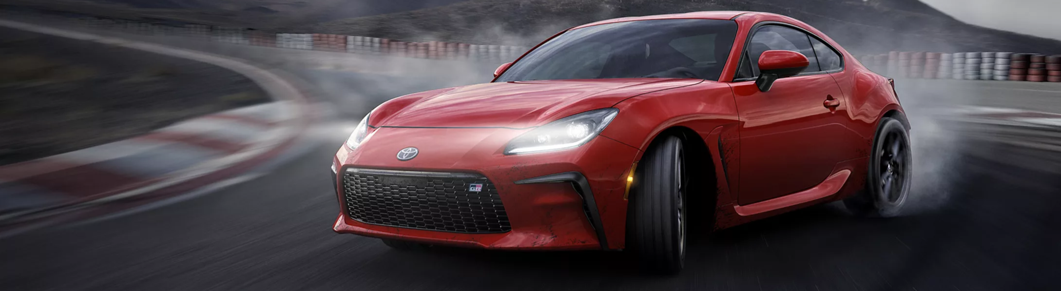 A red 2026 Toyota GR86 sports car blazes around the bend in a racetrack with its high beams on, kicking up clouds of mist from its back tires as it drifts.