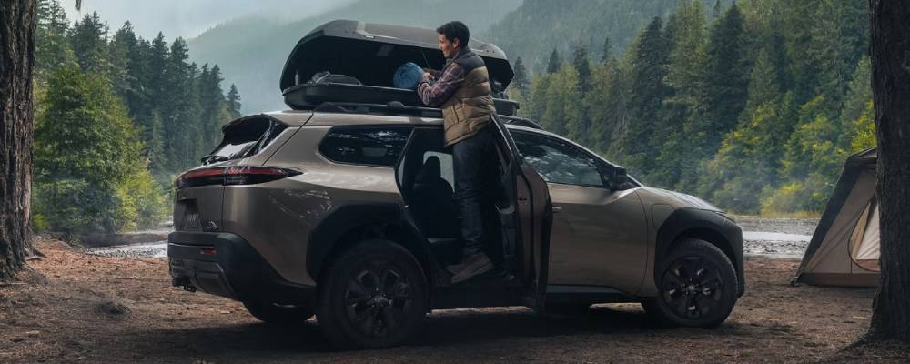 A beige 2026 Toyota bZ Woodland with a roof box. A man packs it up on a woodland campsite. New 2026 Toyota bZ Woodland for sale at Heffner Toyota in Kitchener, ON